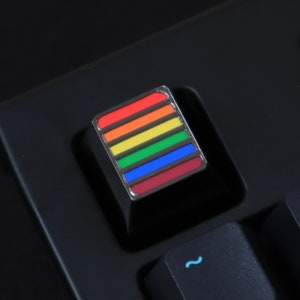 May include: A rainbow-colored keycap for a mechanical keyboard. The keycap is made of plastic and has a silver metal frame. The keycap is designed to look like a rainbow flag.