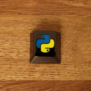 Pode incluir: Uma tecla quadrada cinza escuro com o logotipo Python em azul e amarelo sobre um fundo preto. A tecla est&aacute; sobre uma superf&iacute;cie de madeira.