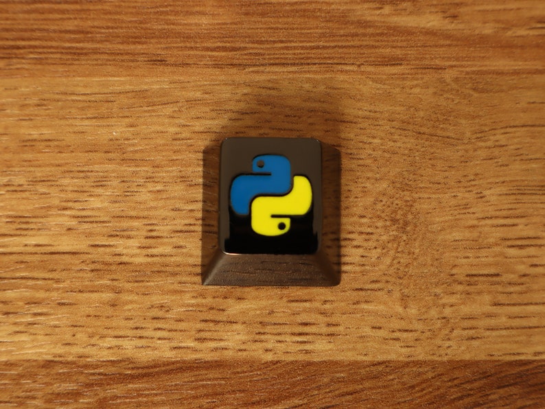 Pode incluir: Uma tecla quadrada cinza escuro com o logotipo Python em azul e amarelo sobre um fundo preto. A tecla est&aacute; sobre uma superf&iacute;cie de madeira.