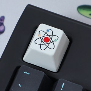 May include: A white keyboard keycap featuring a black atom symbol with colourful dots. The atom has a red centre.