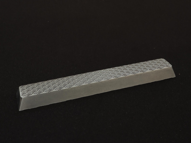 May include: A rectangular, light gray keyboard keycap with a repeating wave pattern in a darker gray. The keycap is angled and appears to be made of a solid material. The background is a solid black.