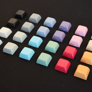 May include: A collection of 18 square keycaps in various colors, including black, gray, light blue, green, pink, red, orange, and yellow. The keycaps are arranged in a triangular pattern on a black surface.