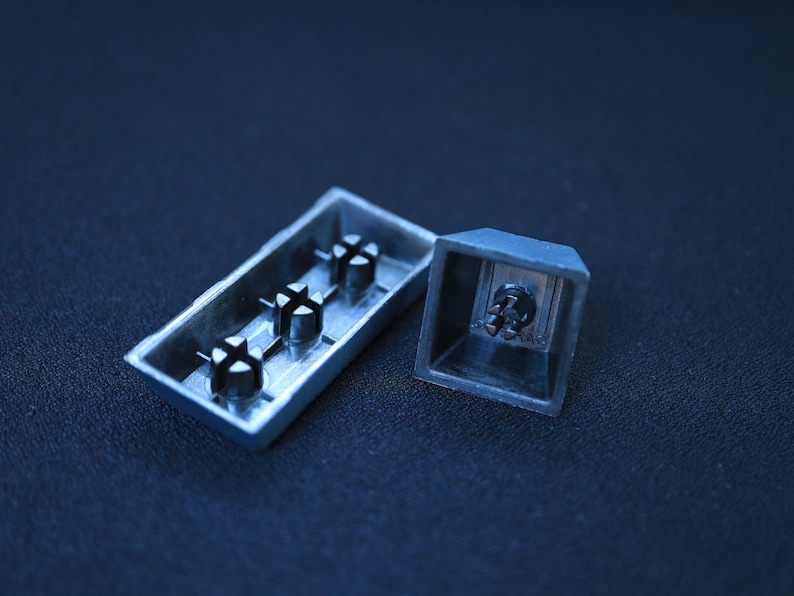 May include: Close-up of two detached, silver-colored keyboard key components. One is a rectangular base with three black posts, and the other is a square keycap. The components are set against a dark blue background.