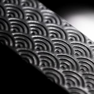 Könnte beinhalten: Nahaufnahme einer strukturierten, dunkelgrauen Oberfläche mit einem sich wiederholenden Wellenmuster. Das Design ist geprägt und erzeugt ein subtiles Relief. Das Bild ist gut beleuchtet und hebt die Details des Musters hervor.