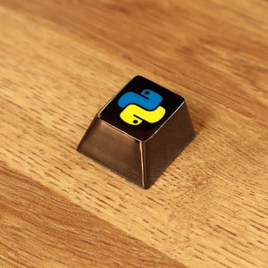 Pode incluir: Uma tecla inclinada marrom escuro com uma parte superior preta com o logotipo da linguagem de programa&ccedil;&atilde;o Python em amarelo e azul. A tecla est&aacute; sobre uma superf&iacute;cie de madeira.
