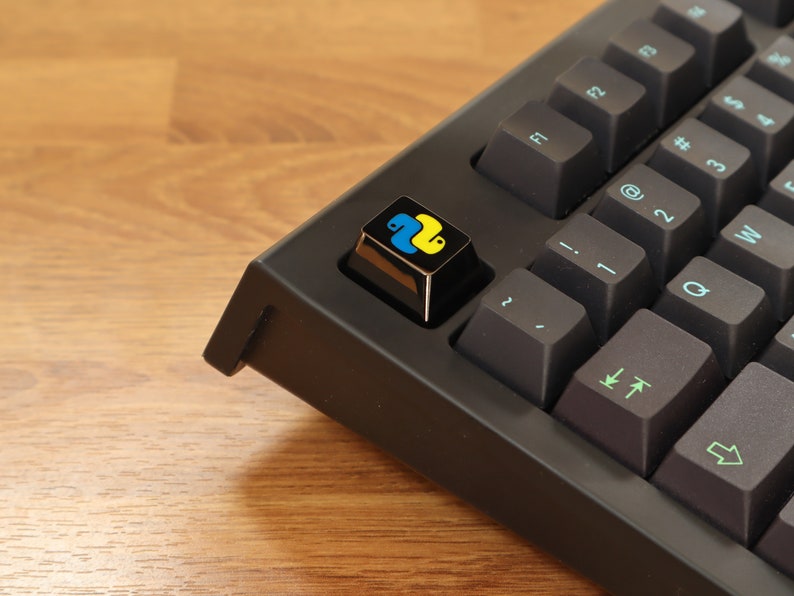 Pode incluir: Close-up de um teclado mec&acirc;nico preto com uma tecla personalizada. A tecla apresenta o logotipo da linguagem de programa&ccedil;&atilde;o Python em amarelo e azul sobre um fundo preto. As outras teclas s&atilde;o cinza escuro com letras turquesa.