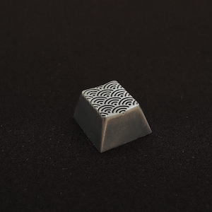May include: A custom keycap with a gray body and a white and black wave pattern on the top surface. The keycap is angled and appears to be made of a solid material. The background is a dark, textured surface.