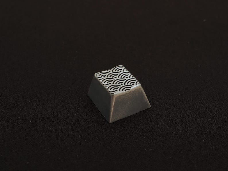 May include: A custom keycap with a gray body and a white and black wave pattern on the top surface. The keycap is angled and appears to be made of a solid material. The background is a dark, textured surface.