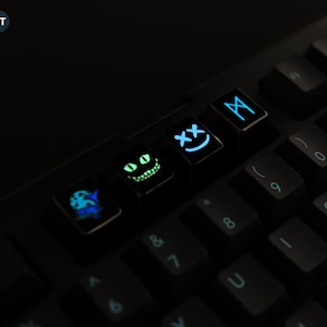 Pode incluir: Close-up de um teclado preto com teclas iluminadas. As teclas apresentam vários designs, incluindo um rosto sorridente, um sorriso do gato de Cheshire e outros símbolos. As teclas brilham com luz azul e verde contra o fundo escuro. A palavra "NIGHT" é visível.