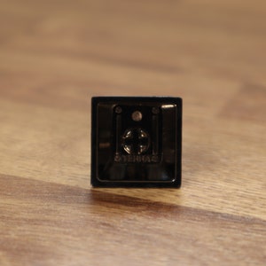 Pode incluir: Um pequeno acess&oacute;rio de c&acirc;mera preto, em forma quadrada, com um design met&aacute;lico em forma de cruz no centro. A palavra "STERHA" est&aacute; impressa na parte inferior. O acess&oacute;rio est&aacute; sobre uma superf&iacute;cie de madeira.