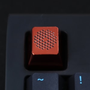May include: A red mechanical keyboard keycap with a black, circular pattern. The keycap is on a black keyboard.