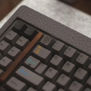 May include: Close-up of a gray mechanical keyboard with gray keycaps. The keyboard has a wooden accent and a keycap with a wave pattern. The keys include Tab, Q, W, E, and R.