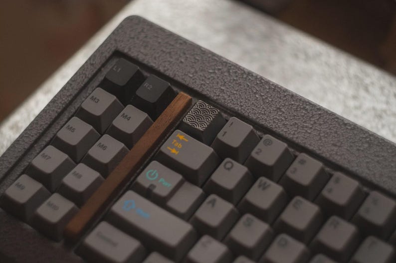 May include: Close-up of a gray mechanical keyboard with gray keycaps. The keyboard has a wooden accent and a keycap with a wave pattern. The keys include Tab, Q, W, E, and R.