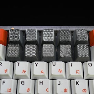 May include: Close-up of a mechanical keyboard with custom keycaps. The keycaps feature various geometric patterns in silver on a dark grey background. Orange function keys are on the sides, and white keys with Japanese characters are in the foreground.