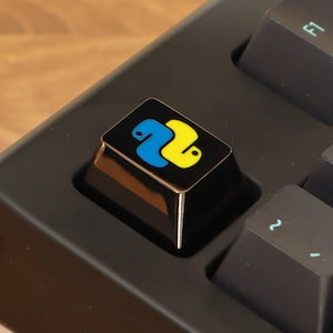 May include: A close-up of a black keyboard with a custom keycap. The keycap displays the Python logo, a blue and yellow snake design, on a black background. Other keys are black with light blue symbols.