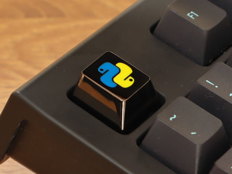 Pode incluir: Close-up de um teclado preto com uma tecla personalizada. A tecla apresenta o logotipo Python, um design de cobra azul e amarelo, em um fundo preto. Outras teclas s&atilde;o pretas com s&iacute;mbolos azul claro.