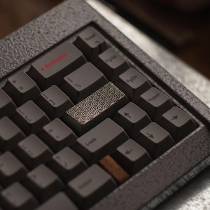 May include: Close-up of a custom mechanical keyboard with dark gray keycaps and a textured gray frame. The Backspace key is red. The space bar has a decorative pattern. A wooden key is visible.