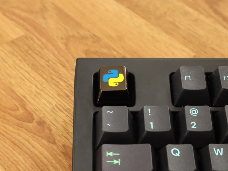 Pode incluir: Close-up de um teclado mec&acirc;nico preto com uma tecla personalizada com o logotipo da linguagem de programa&ccedil;&atilde;o Python. A tecla &eacute; met&aacute;lica com um &iacute;cone de cobra azul e amarelo. O teclado est&aacute; sobre uma superf&iacute;cie de madeira.