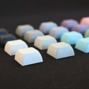 May include: A row of colorful keycaps in various shades of blue, white, and gray. The keycaps are arranged in a gradient pattern, starting with the darkest blue on the left and transitioning to the lightest gray on the right.