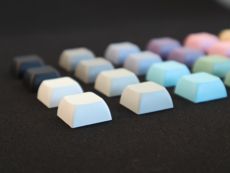 May include: A row of colorful keycaps in various shades of blue, white, and gray. The keycaps are arranged in a gradient pattern, starting with the darkest blue on the left and transitioning to the lightest gray on the right.
