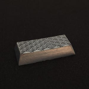 May include: A rectangular keycap with a Japanese wave pattern in black and white. The bottom half of the keycap has a metallic silver finish. The keycap is angled and sits on a black surface.