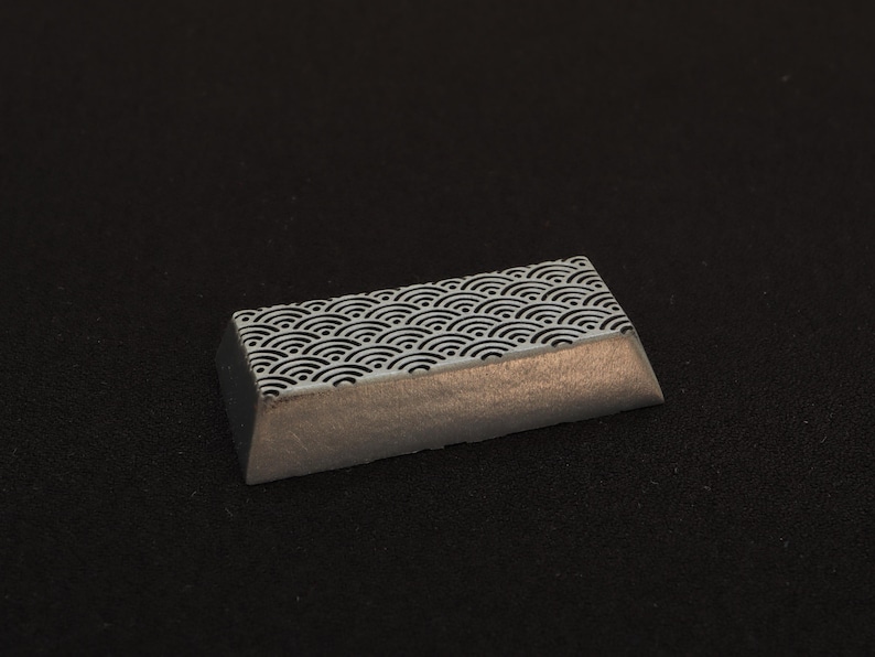 May include: A rectangular keycap with a Japanese wave pattern in black and white. The bottom half of the keycap has a metallic silver finish. The keycap is angled and sits on a black surface.