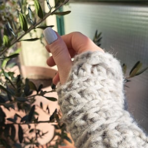 Puede incluir: Primer plano de una mano con un calientamanos de punto gris claro. El calientamanos está hecho de un hilo suave y esponjoso y tiene un diseño simple y texturizado.