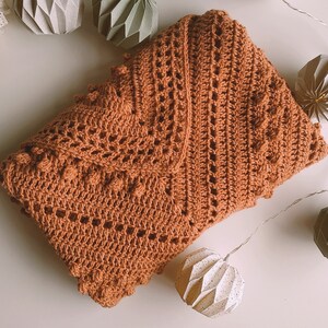 Puede incluir: Una manta de ganchillo de color marrón cálido con un patrón de encaje y pequeños bultos en relieve. La manta está doblada por la mitad y está sobre una superficie blanca.