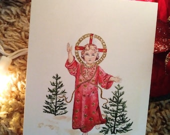 May include: A vintage Christmas card featuring a watercolor illustration of a young child in a red robe with gold accents, standing between two small evergreen trees. The child is wearing a halo with a cross and the word "JHS" is visible on the robe.