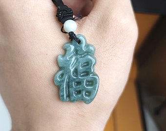 Puede incluir: Un colgante de jade tallado con caracteres chinos en un cordón negro con una cuenta blanca. El colgante es verde y tiene un acabado brillante.