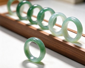 May include: A collection of smooth, polished jade rings in varying shades of translucent green. Six rings are arranged in a diagonal line, while one single ring stands upright in the foreground, showcasing a glossy, rounded band design.