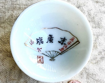 Może przedstawiać: Biała ceramiczna filiżanka do sake z wzorem złotego i czarnego wachlarza i japońskimi znakami. Filiżanka jest mniej więcej wielkości polskiego grosza.