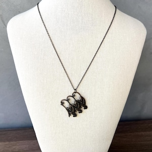 Puede incluir: Un collar de color bronce oscuro con un colgante que presenta tres siluetas de pájaros estilizadas. El collar se exhibe sobre un busto de maniquí blanco sobre un fondo neutro. La cadena es delicada y el colgante tiene un diseño detallado.