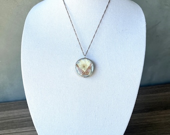 May include: A round pendant necklace with a dried flower and fern preserved in clear resin. The pendant is suspended from a delicate, twisted metal chain. The necklace is displayed on a white mannequin.