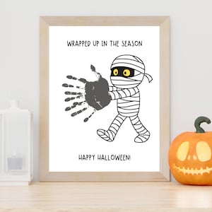 K&ouml;nnte beinhalten: Ein gerahmter Druck mit einer Cartoon-Mumie mit gelben Augen, einem grauen Handabdruck und dem Text WRAPPED UP IN THE SEASON und HAPPY HALLOWEEN! Die Mumie ist in schwarz-wei&szlig;er Strichzeichnung dargestellt, wie sie geht und die Hand ausstreckt.