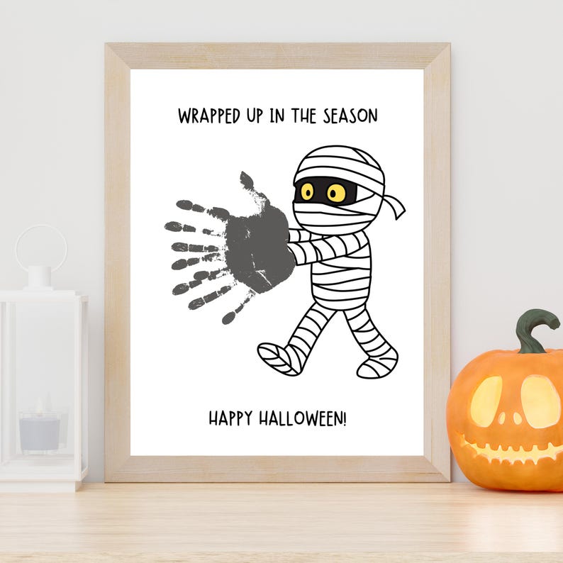 K&ouml;nnte beinhalten: Ein gerahmter Druck mit einer Cartoon-Mumie mit gelben Augen, einem grauen Handabdruck und dem Text WRAPPED UP IN THE SEASON und HAPPY HALLOWEEN! Die Mumie ist in schwarz-wei&szlig;er Strichzeichnung dargestellt, wie sie geht und die Hand ausstreckt.