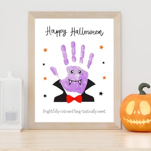 K&ouml;nnte beinhalten: Ein gerahmter Druck mit einem violetten Handabdruck, der als Vampir mit schwarzem Umhang, roter Fliege und wei&szlig;en Fangz&auml;hnen gestaltet ist. Der Text lautet Happy Halloween und Frightfully cute and fang-tastically sweet, umgeben von kleinen Sternen.