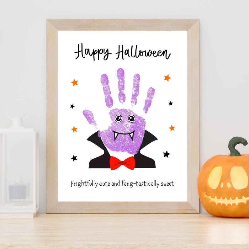 K&ouml;nnte beinhalten: Ein gerahmter Druck mit einem violetten Handabdruck, der als Vampir mit schwarzem Umhang, roter Fliege und wei&szlig;en Fangz&auml;hnen gestaltet ist. Der Text lautet Happy Halloween und Frightfully cute and fang-tastically sweet, umgeben von kleinen Sternen.