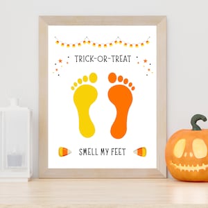 K&ouml;nnte beinhalten: Ein gerahmter Druck mit dem Text TRICK-OR-TREAT und SMELL MY FEET in schwarzer Schrift. Das Design zeigt einen gelben Fu&szlig;abdruck, einen orangen Fu&szlig;abdruck, eine Zuckermais-Girlande, kleine orange Sterne und zwei Zuckermais-Illustrationen.
