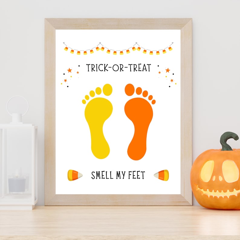 K&ouml;nnte beinhalten: Ein gerahmter Druck mit dem Text TRICK-OR-TREAT und SMELL MY FEET in schwarzer Schrift. Das Design zeigt einen gelben Fu&szlig;abdruck, einen orangen Fu&szlig;abdruck, eine Zuckermais-Girlande, kleine orange Sterne und zwei Zuckermais-Illustrationen.