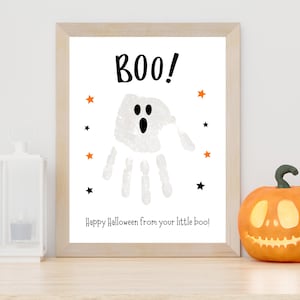 K&ouml;nnte beinhalten: Ein gerahmter Druck mit einem wei&szlig;en Handabdruck-Gespenst mit schwarzen Augen und Mund, dem Text BOO! oben und dem Text Happy Halloween from your little boo! unten, akzentuiert durch kleine orangefarbene und schwarze Sterne auf wei&szlig;em Grund.