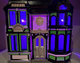 May include: A decorative haunted house with a black roof, black and green trim, and purple and white walls. The windows glow with a bright blue light. The front door is silver with a decorative oval above it. A spooky Halloween decoration.