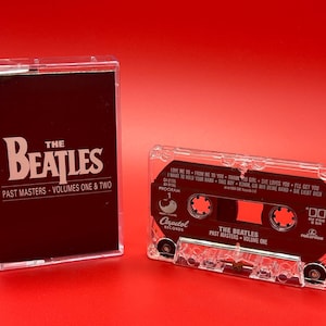 May include: A clear plastic cassette tape case with a red and white label. The label says "The Beatles Past Masters - Volume One" and has the Capitol Records logo. The cassette tape is in the case and has a white label with the song titles "Love Me Do", "From Me To You", "Thank You Girl", "She Loves You", "I'll Get You", "I Want To Hold Your Hand", "This Boy", "Komm, Gib Mir Deine Hand", "She's Left Me Doch".