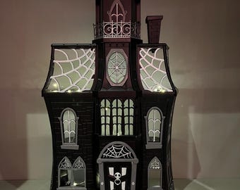 May include: A black and gray Halloween haunted house decoration with a bat on top. The house features spiderwebs, arched windows, and a skull on the front door. Two orange pumpkins sit on the steps. The house is approximately 18 inches tall.