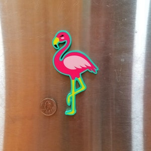 May include: Pink flamingo magnet with yellow legs and a turquoise outline. The magnet is on a stainless steel surface.
