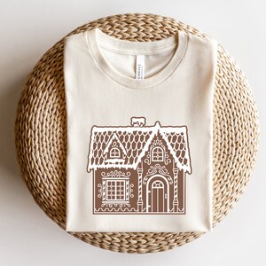 May include: A beige t-shirt with a brown gingerbread house design. The house has white icing details and a snowy roof.
