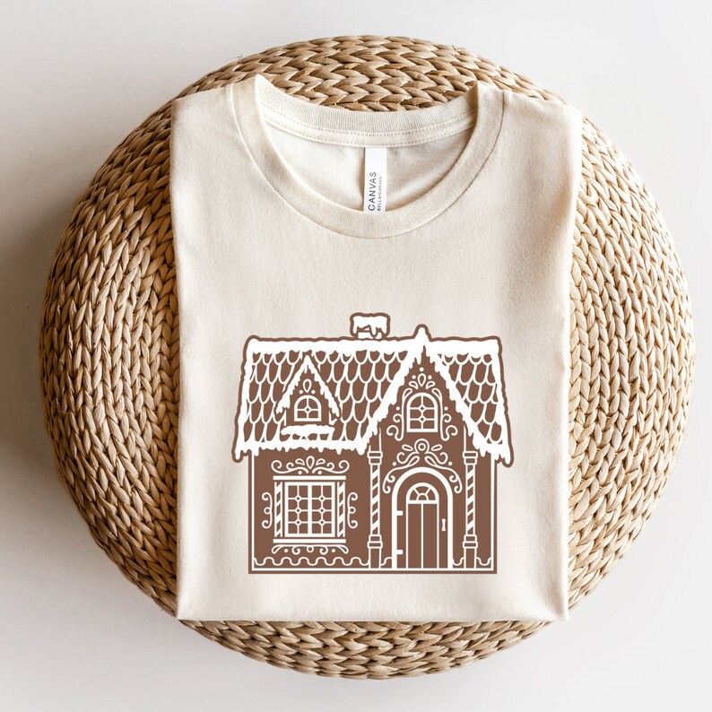 May include: A beige t-shirt with a brown gingerbread house design. The house has white icing details and a snowy roof.