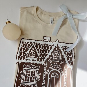 May include: A beige t-shirt with a white gingerbread house graphic. The house has brown trim and white icing details.