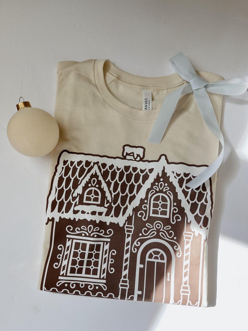 May include: A beige t-shirt with a white gingerbread house graphic. The house has brown trim and white icing details.