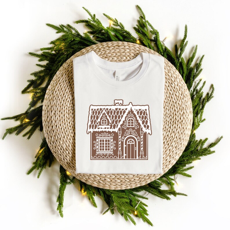 May include: White t-shirt with a brown gingerbread house design. The house has a white roof and a brown door.